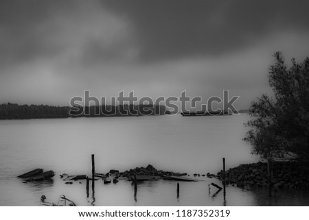 Monochrome picture.
On a calm river a ship is in the background.
Rocks and wood in the foreground.