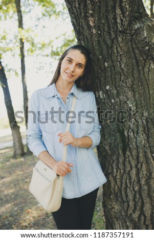 young girl student walks in the autumn city Park after school. street style clothing: denim shirt and stylish shoulder bag. long hair and problem skin: pimples and blemishes