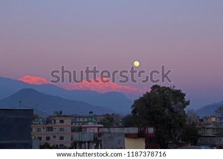 At dusk, a full moon rises above snowy peaks of the himalayas in Nepal, with buildings in the foreground. The last light lets the mountains shine red and pink. The picture was taken in Pokhara.