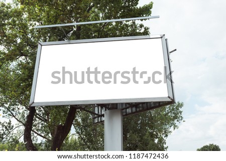 Blank White and Isolated Mockup. Billboard for Poster or Canvas standing on the sidewalk in the city among green trees in nice weather. Bottom view for a large advertisement.