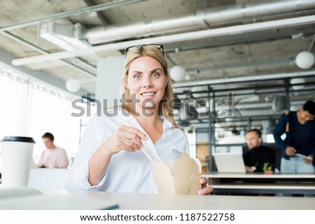 Portrait of cheerful blonde woman eating Chinese food at workplace and smiling at camera, copy space