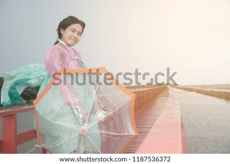 Asian woman pose of standing in Korean Hanbok dresses with transparent umbrella on red wooden bridge.