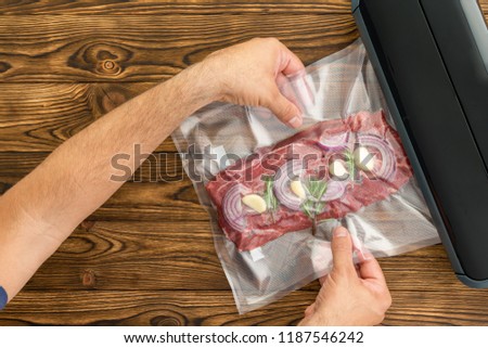 Unidentified adult man sliding plastic packaged meat with onion and garlic into black machine to preserve meat Royalty-Free Stock Photo #1187546242
