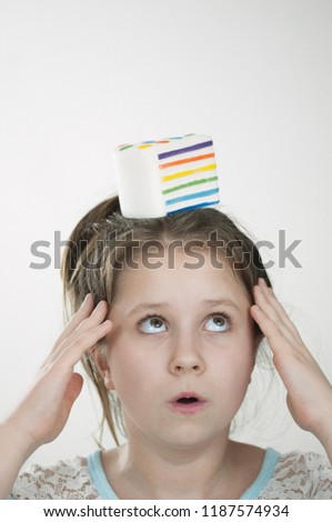 The vertical picture of a girl with a sguishy toy cake on her head and with a hands close to her temples and looking at cake over light background