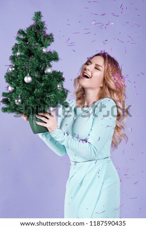 A charming blonde in a mint dress holds a small Christmas tree in her hands and laughs, sparkles and confetti are pouring from above.