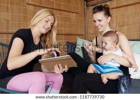 Attractive young woman holding tablet and showing cartoons to adorable little son of her friend while waiting for yoga practice in studio