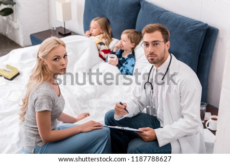 high angle view of pediatrician and mother looking at camera while sick kids lying in bed on background