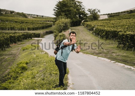 
Young and adventurous man hitchhiking on a side road of France surrounded by vineyards on a cloudy day. Travel photography. Lifestyle