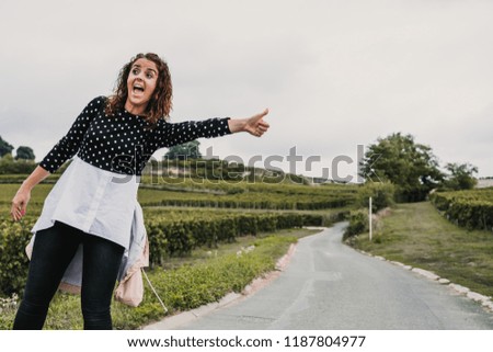 
Young and adventurous woman hitchhiking on a side road of France surrounded by vineyards on a cloudy day. Travel photography. Lifestyle