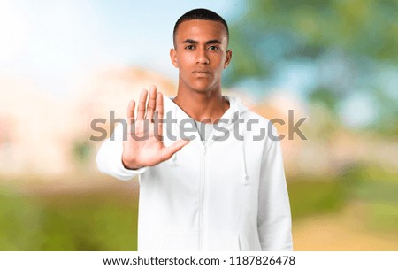Dark-skinned young man with white sweatshirt making stop gesture with her hand denying a situation that thinks wrong in a park