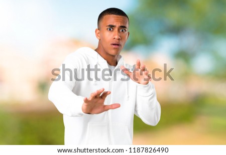 Dark-skinned young man with white sweatshirt is a little bit nervous and scared stretching hands to the front in a park