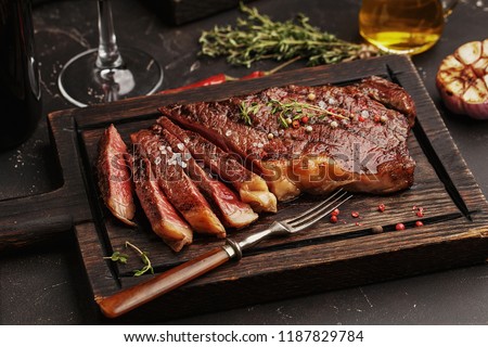 Medium rare sliced grilled striploin beef steak served on wooden board with vintage fork, glass of wine, herbs and spices Royalty-Free Stock Photo #1187829784