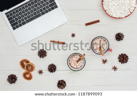 flat lay with laptop, mugs of hot chocolate and cinnamon sticks with marshmallows on wooden table 