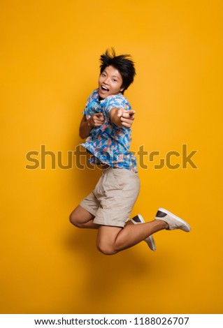 Image of happy emotional young asian man jumping isolated over yellow background pointing.