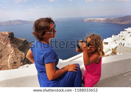 a little girl takes pictures of her mother against the beautiful sea view of Santorini, Greece