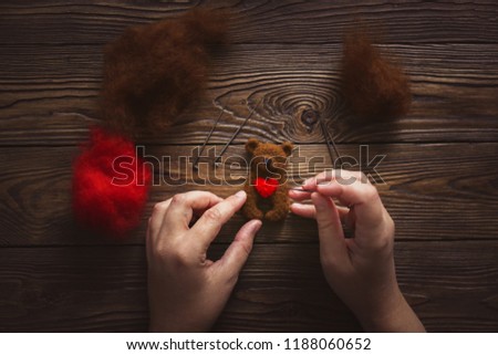 Dry felting, women's hands with needles are laying heart on the chest of a bear made of wool. Wooden brown background