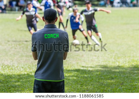 Male soccer or football coach in gray shirt with word COACH written on back, standing on the sideline watching his team play, good for sport or coaching concept Royalty-Free Stock Photo #1188061585