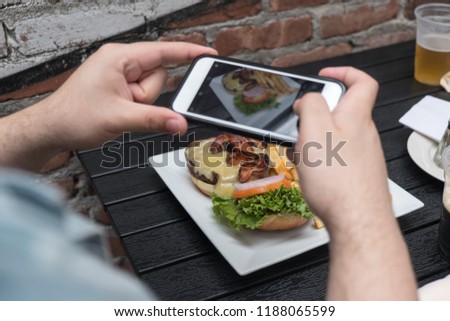 Close up, man takes photo of food with mobile phone at an outdoor bar.  Taking a picture of your food with your phone. Hamburger, fries, and beer on white plate outside on a black table focus on food