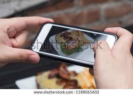 Man takes photo of food with mobile phone at an outdoor bar.  Taking a picture of your food with your phone. Hamburger, fries, and beer on a white plate outside on a black table focus on cell phone.