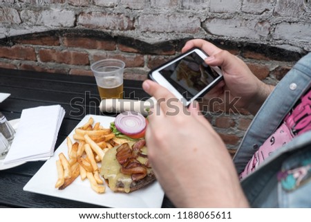 Man holding a mobile phone taking a photo of his food. Smartphone food photography. Taking a picture of hamburger, beer, and french fries at an outdoor bar with a mobile phone.