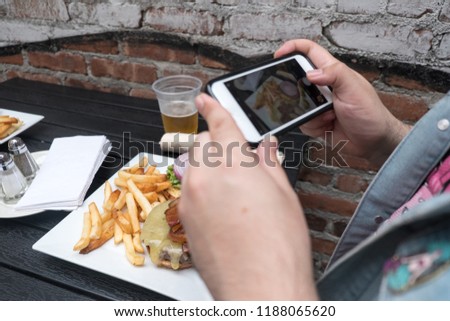 Man holding a mobile phone taking a photo of his food. Smartphone food photography. Taking a picture of hamburger, beer, and french fries at an outdoor bar with a cell phone.