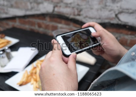 Man holding a mobile phone taking a photo of his food. Smartphone food photography. Taking a picture of hamburger and french fries at an outdoor bar with a smartphone.