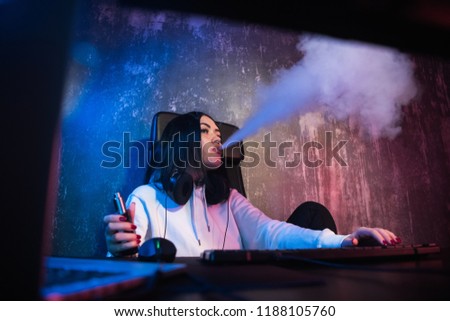 Young brunette woman wearing hoodie, sitting down by desk with computer while smoking e-cigarette