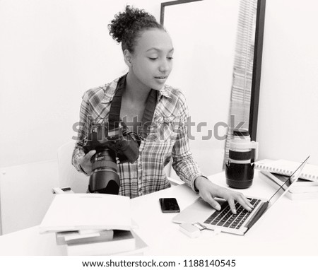 Black and white portrait of a beautiful african american teenager media student holding a camera, learning photography indoors. Young woman using professional equipment and technology.