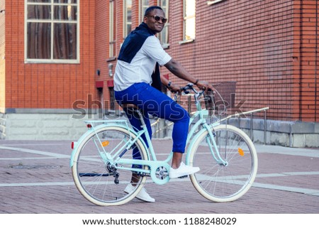handsome African American man stylish well-dressed standing with a blue bicycle red brick building background . sports ecological transport concept