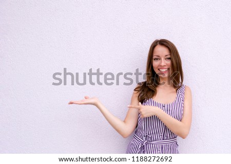 Waist-up portrait of brunette girl presenting your product on palm of hand. Cheerful pretty young woman in bright summer dress on white wall. Copy space in left side. Advertisement concept
