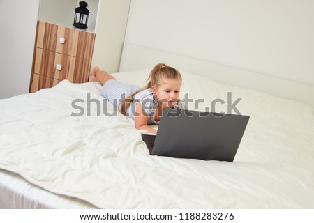 Little blonde girl looking at laptop while lying on bed in her room
