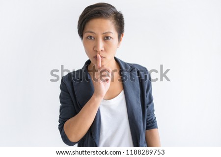 Serious Asian woman making silence gesture and looking at camera. Lady touching mouth with forefinger. Secret concept. Isolated front view on white background.