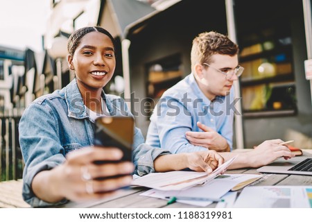 Cheerful African American woman taking picture on mobile phone of working process with male caucasian colleague,positive hipster girl using smartphone camera for photographing exam preparation process
