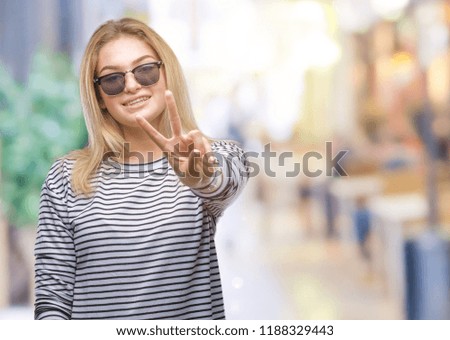 Young caucasian woman wearing sunglasses over isolated background smiling with happy face winking at the camera doing victory sign. Number two.