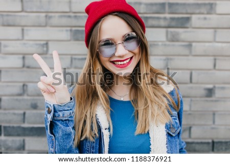 Charming european woman in casual clothes posing with cute smile and peace sign. Outdoor shot of elegant laughing girl in red hat.