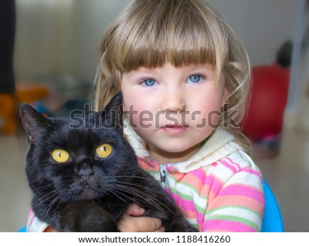 A beautiful little girl with blue eyes is holding a black cat. Friendship with pets. Close-up.