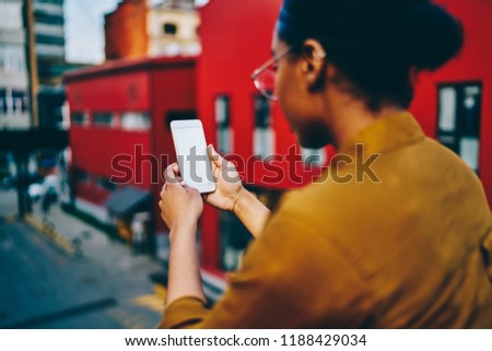 Selective focus on modern smartphone with mock up screen, rear view of dark skinned hipster girl holding mobile phone connected to 4G for chatting and blogging in social networks. Copy space screen