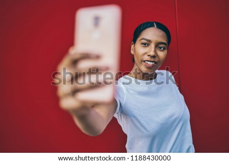 Selective focus on attractive dark skinned young woman posing for selfie on red wall background, cheerful African American hipster girl holding modern smartphone for taking images for share in blog