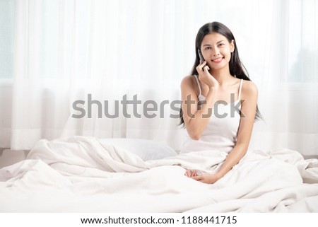 Woman talking on phone in bed. Asian woman in white top, talking, happy mood. Modern living concept.