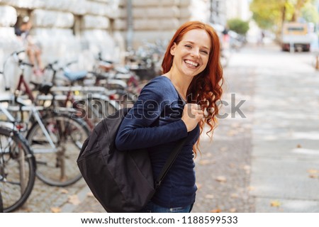 Attractive young redhead woman wearing a backpack leaving a bicycle rank in town turning to give the camera a lovely vivacious smile