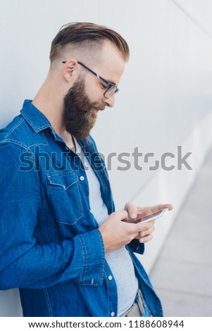 Casual bearded man leaning on an exterior white wall texting on his mobile phone in a close up side view