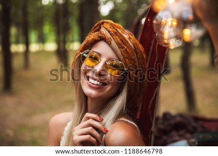 Photo of blond traveler woman wearing stylish accessories smiling while resting in forest camp