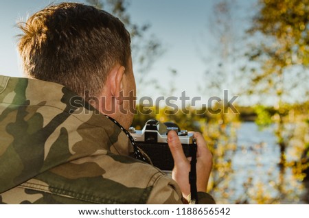 A photographer dressed in camouflage jacket with film camera in his hands takes pictures on the shore of a lake or river. Closeup back view.