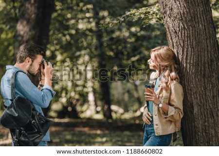 side view of man taking picture of smiling girlfriend with coffee to go in park