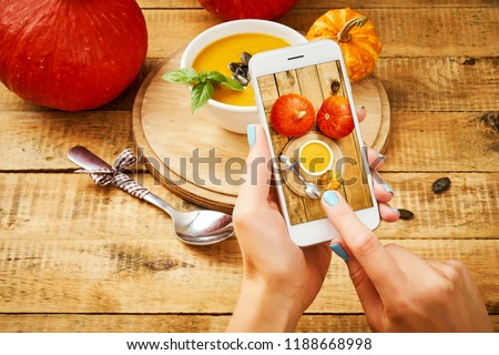woman's hands are holding the phone and taking a picture of a delicious pumpkin soup on a wooden table