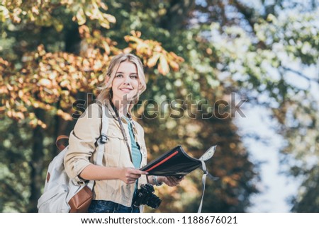 portrait of young smiling traveler with backpack, photo camera and map in park