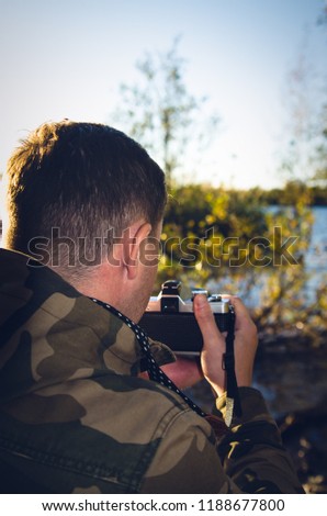 A photographer dressed in camouflage jacket with film camera in his hands takes pictures on the shore of a lake or river. Back view with darkening effect.