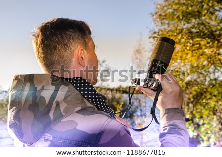 A photographer dressed in camouflage jacket with a vintage film camera in his hands chooses the next frame. Back view with blue and yellow light leaks.