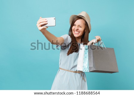 Portrait smiling woman in summer dress straw hat holding packages bags with purchases after shopping doing selfie shot on mobile phone isolated on blue pastel background. Copy space for advertisement