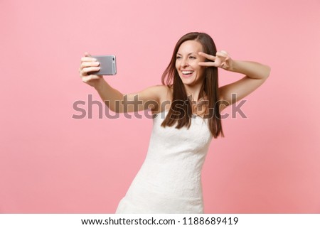 Portrait of joyful bride woman in elegant white wedding dress showing victory sign doing taking selfie shot on mobile phone isolated on pastel pink background. Wedding celebration concept. Copy space